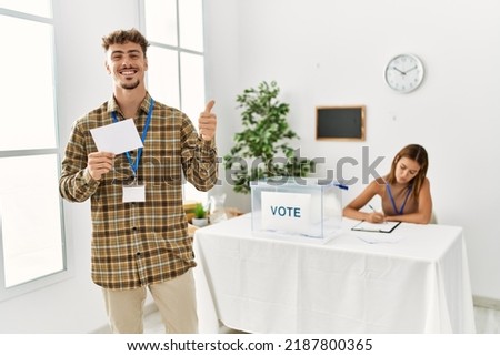 Young handsome man voting putting envelop in ballot box smiling happy and positive, thumb up doing excellent and approval sign 