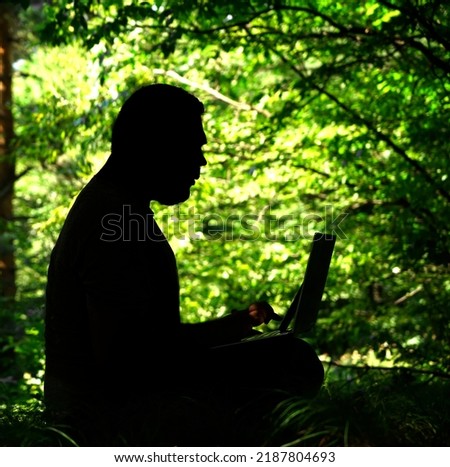 silhouette of middle age  freelancer man is working on laptop on summer forest background. A young business man sits on fresh grass and works on wireless computer and mobile phone. summertime season
