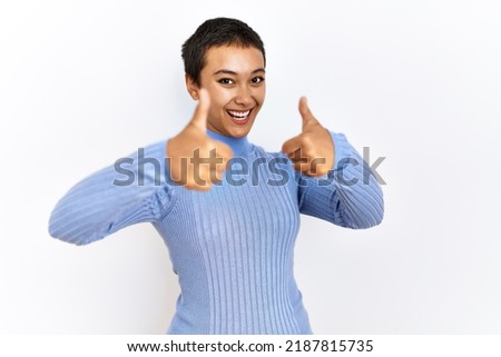 Young hispanic woman with short hair standing over isolated background approving doing positive gesture with hand, thumbs up smiling and happy for success. winner gesture. 