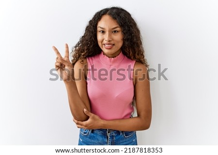 Beautiful hispanic woman standing over isolated background smiling with happy face winking at the camera doing victory sign. number two. 