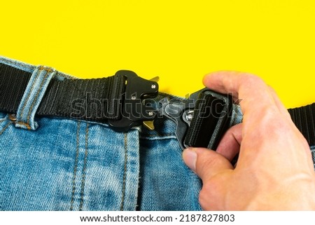A man buttoned a black cloth belt on jeans. Modern men's waistband with dark matted metal buckle.