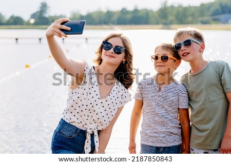 Happy children with teenager girl taking selfie by smartphone at nature. Cute different age boys and girl in eyeglasses having fun together. Vacation, relax, active lifestyle concept.