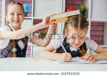 Two girls are sitting at one desk in the classroom. One girl hits another on the head. The problem of bullying at school and the aggression of children concept. Royalty-Free Stock Photo #2187853035