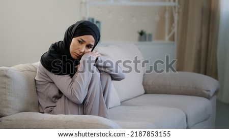 Anxious sad young woman in black hijab sitting on couch in living room suffering from psychological problem trouble at home frustrated muslim girl feeling stress depressed lost in negative thoughts Royalty-Free Stock Photo #2187853155