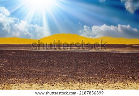 Beautiful wide vast arid dry moroccan desert landscape, black scorched earth, yellow sand dunes, blue dramamtic sky clouds, sun rays backlight - Sahara, Erg Chebbi, Morocco  Royalty-Free Stock Photo #2187856399