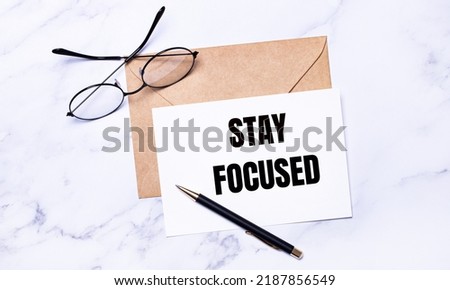 On a light marble background, a craft envelope, glasses, a pen and a white card with the text STAY FOCUSED