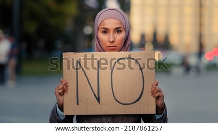 Stop racism No concept Arab immigrant Muslim woman in hijab protests against discrimination vax vaccination standing in city. Islamic girl holding cardboard slogan banner with text no disagree Royalty-Free Stock Photo #2187861257