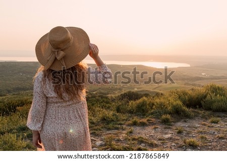 Woman holds hat in her hand and watches sunset over lake. Selective focus. Picture for articles about women, success, psychology.