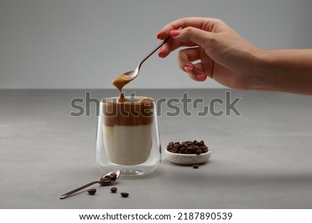 Cup with Dalgona coffee. Iced fluffy creamy whipped trend drink with coffee foam and milk in a glass with double glass. Female hand takes foam from a cup of coffee with a spoon. Shallow depth of field Royalty-Free Stock Photo #2187890539