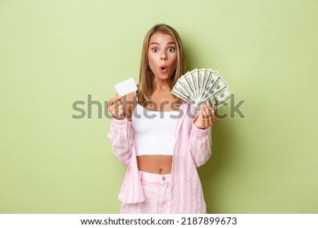 Image of impressed and excited blond woman, holding cash and credit card, standing over green background