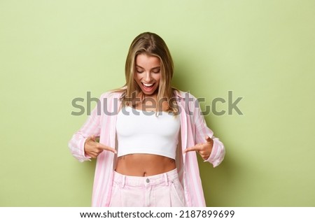 Image of stylish modern girl with blond hair, wearing cropped top with pink shirt, pointing fingers at flat belly and looking pleased, losing weight, standing over green background Royalty-Free Stock Photo #2187899699