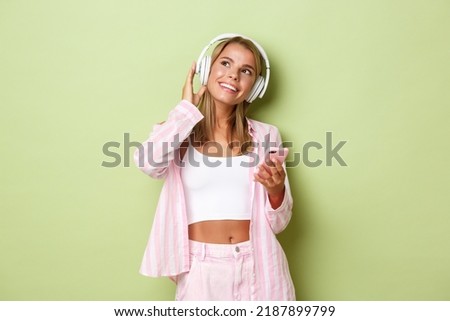 Image of stylish glamour girl with blond short hair, looking at upper left corner dreamy while listening to music in wireless headphones, holding mobile phone, green background