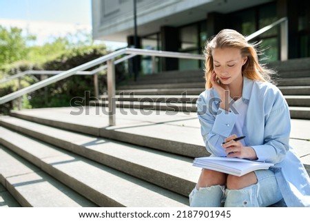 Pretty blonde girl college student holding notebook sitting on stairs outside campus writing checklist essay for university study course, looking for inspiration, thinking of new creative ideas plan Royalty-Free Stock Photo #2187901945