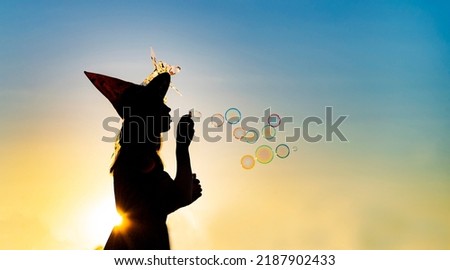 Black silhouette of witch blowing soap bubbles isolated on fairy sunset sky background.Beautiful young woman in black robe and wizard hat conjuring, making magic. Halloween party art design.Copy space