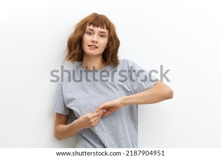 a sweet, beautiful, red-haired, curly-haired woman stands on a white background in a gray cotton T-shirt and holds her hands in a relaxed gesture. Horizontal studio photo with an empty space for