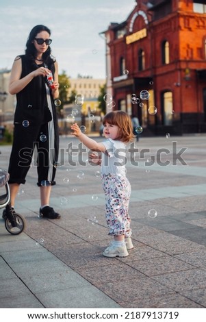 little girl playing with her mother in soap blowing bubbles on the street in summer