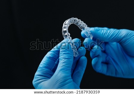 Invisible orthodontics cosmetic aligners in hands of doctor wearing sterile gloves isolated on a black background, tooth aligners, plastic braces. Modern teeth retainers created on a 3d printer. Royalty-Free Stock Photo #2187930359