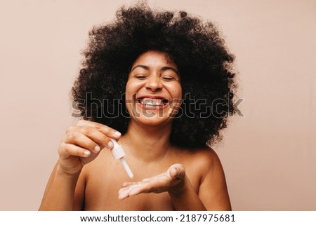 Gorgeous young woman smiling cheerfully while dropping cosmetic face serum onto her palm. Happy woman with Afro hair treating her skin with a moisturizing beauty product. Royalty-Free Stock Photo #2187975681