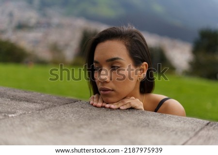 face of a young Latin brunette woman with short hair leaning against a platform worried and sad, mountain and city background, beauty and emotions