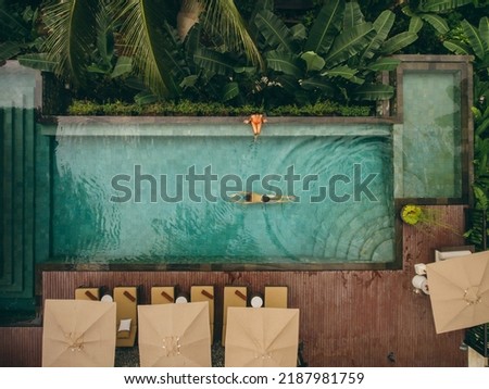Aerial shot of woman sitting at the poolside with man swimming in pool. Couple enjoying holiday at luxury resort.