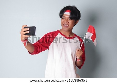 Portrait of attractive Asian man in t-shirt with red and white ribbon on head, taking a picture of himself while holding Indonesia flag. Isolated image on gray background