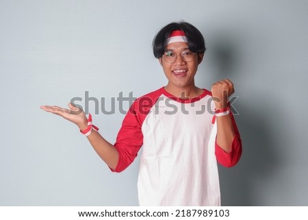 Portrait of attractive Asian man in t-shirt with red and white ribbon on head, raising his fist while demonstrating product. Isolated image on gray background