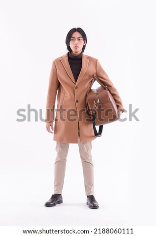 fashion model. full body Young man with hairstyle in brown coat ,black sweater holding handbag  posing on white background

