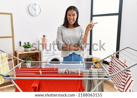 Young hispanic woman putting fresh laundry on clothesline smiling with happy face winking at the camera doing victory sign. number two. 