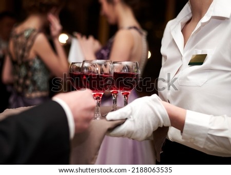 Close-up picture of waiter's hands wearing white gloves holding a tray with red wine, serving alcohol drinks. Catering service at special occasion, event. Hospitality industry concept.