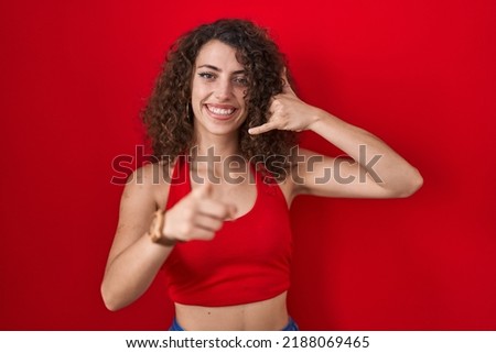 Hispanic woman with curly hair standing over red background smiling doing talking on the telephone gesture and pointing to you. call me. 