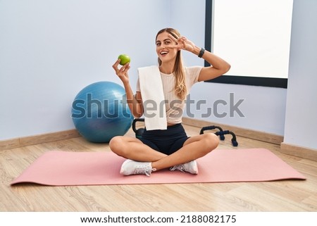 Young blonde woman wearing sportswear and towel holding healthy apple doing peace symbol with fingers over face, smiling cheerful showing victory 