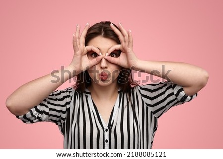 Funny young female with long dark hair in striped shirt pouting lips and making binoculars gesture against pink background