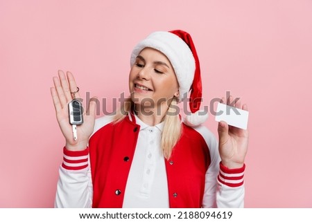 Smiling blonde woman in santa hat holding car key and empty driving license isolated on pink