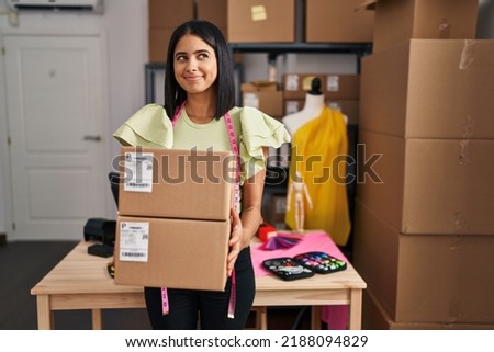 Young hispanic woman holding delivery boxes at small business smiling looking to the side and staring away thinking. 