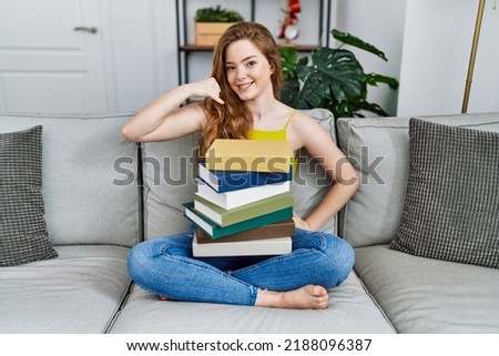 Young caucasian woman sitting on the sofa with books at home smiling doing phone gesture with hand and fingers like talking on the telephone. communicating concepts. 
