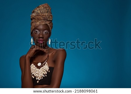 Serious elegant black girl looking at camera. Beautiful young slim woman wearing traditional african outfit and accessories. Female beauty. Isolated on blue background. Studio shoot. Copy space
