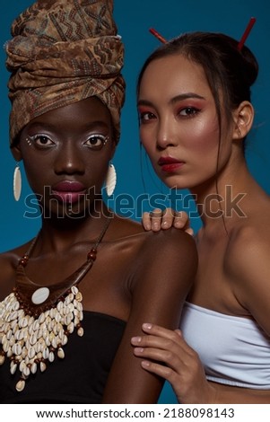 Cropped of attractive multiethnic girl friends in their national traditional styles looking at camera. Black girl wearing african outfit and accessories with asian woman. Blue background. Studio