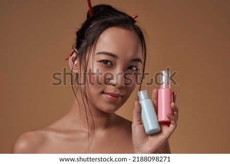 Elegant asian girl holding two colorful nail polishes and looking at camera. Beautiful young brunette woman wearing tank top. Female beauty. Isolated on orange background. Studio shoot. Copy space