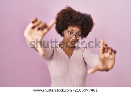 Young african american woman standing over pink background doing frame using hands palms and fingers, camera perspective 