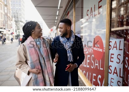 Cheerful couple arm in arm walking past sale signs in store window in town