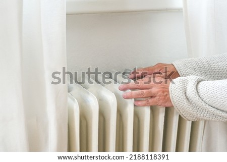 Hands of an elderly woman in woolen clothes feeling low temperature on an old heater, suffering from inflation and rising energy prices, copy space, selected focus, narrow depth of field Royalty-Free Stock Photo #2188118391
