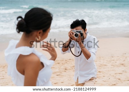Asian couple enjoying beach vacation holiday. Boyfriend photographer taking a picture of his girlfriend by seaside in summer. A couple wearing a white shirt and dress. Royalty-Free Stock Photo #2188124999