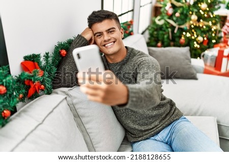 Young hispanic man make selfie by the smartphone sitting on sofa by christmas tree at home