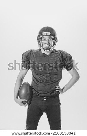 Portrait of young smiling man, american football player posing in full equipment with ball. Black and white photography. Concept of sport, retro style, 20s, fashion, action, college sport, youth