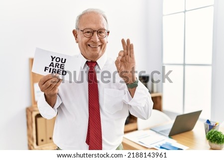 Senior business man holding you are fired banner at the office doing ok sign with fingers, smiling friendly gesturing excellent symbol 