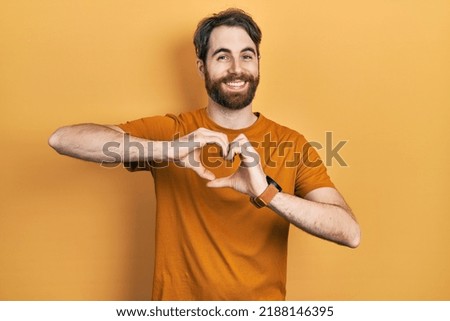 Caucasian man with beard wearing casual yellow t shirt smiling in love doing heart symbol shape with hands. romantic concept. 