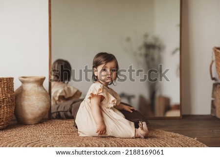 Pretty toddler girl in beige dress and shoes sitting on eco rug in the room with big mirror. Royalty-Free Stock Photo #2188169061