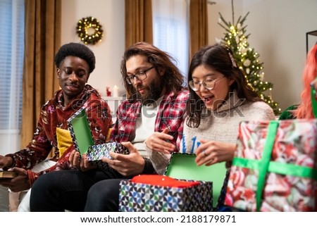 African-looking guy holds book in hands and discusses what he read with friend. Friends are sitting on couch and next to them is smiling girl of Asian appearance, who is unpacking Christmas present.