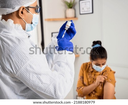  Indian male or man Doctor preparing a dose of vaccination to inject in the arm of a young Asian female wearing protective masks at modern clinic or hospital amid corona virus or COVID 19 epidemic 
