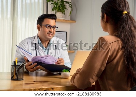 Asian Indian male physician or doctor wearing white apron and stethoscope holding a file of medical report is consulting or prescribing medicines to a Young Female patient in modern clinic or hospital Royalty-Free Stock Photo #2188180983